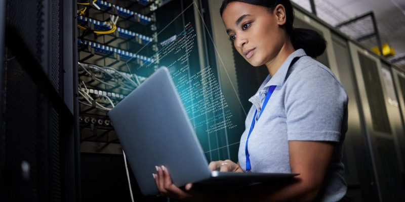 IT-Spezialistin arbeitet an einem Laptop in einem modernen Rechenzentrum mit vernetzten Servern – Symbol für digitale Infrastruktur und Netzwerkmanagement.